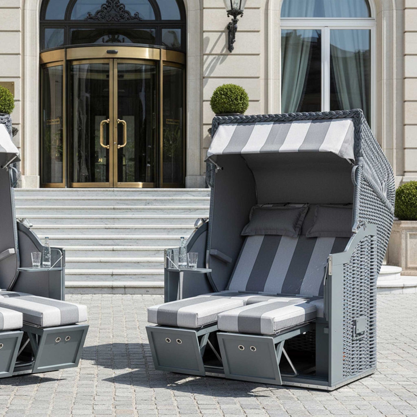 Zwei Strandkorb Sonnenpartner Modelle Classic PE-Geflecht in Onyx Farbe, Holz grau, vor einem eleganten Gebäude.