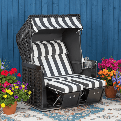 Strandkorb Sonnenpartner Konsul schwarz mit Streifenpolster Dessin 83 vor blauer Wand, umgeben von bunten Blumen